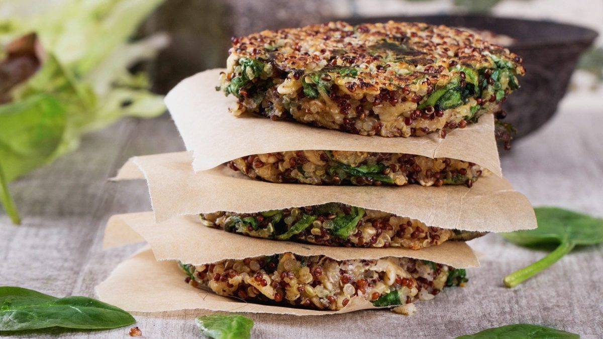 Drei geschichterte Quinoa-Burger-Patties auf einem Untergrund. Zwischen den Patties liegen Backpapierzuschnitte, damit sie nicht aneinander haften. Um die Patties herum liegen Spinatblätter.
