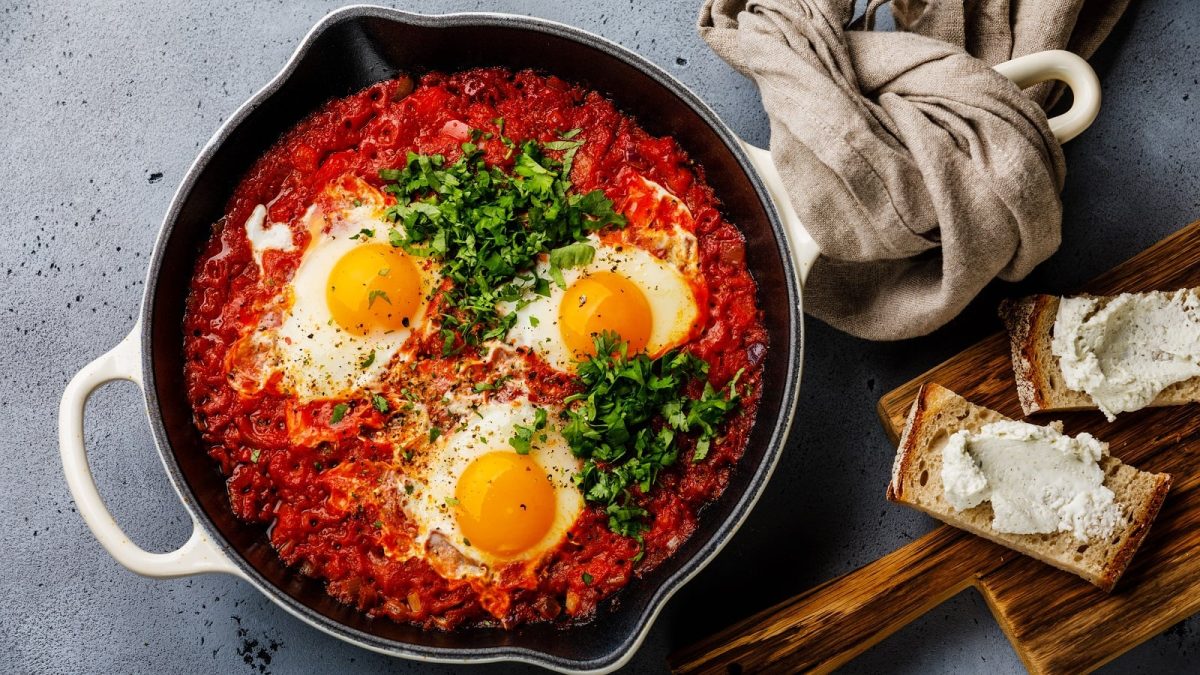 Eine große Pfanne mit Shakshuka mit drei Eiern und viel Koriander und Petersilie garniert neben einem Holzbrett mit mit Frischkäse bestrichenem Brot vor einem grauem Hintergrund, von oben.