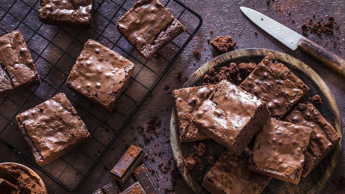 Ein Kuchengitter und rundes Holzbrett mit Süßkartoffel-Brownies in der Draufsicht. Daneben liegen ein Messer und Schokolade.