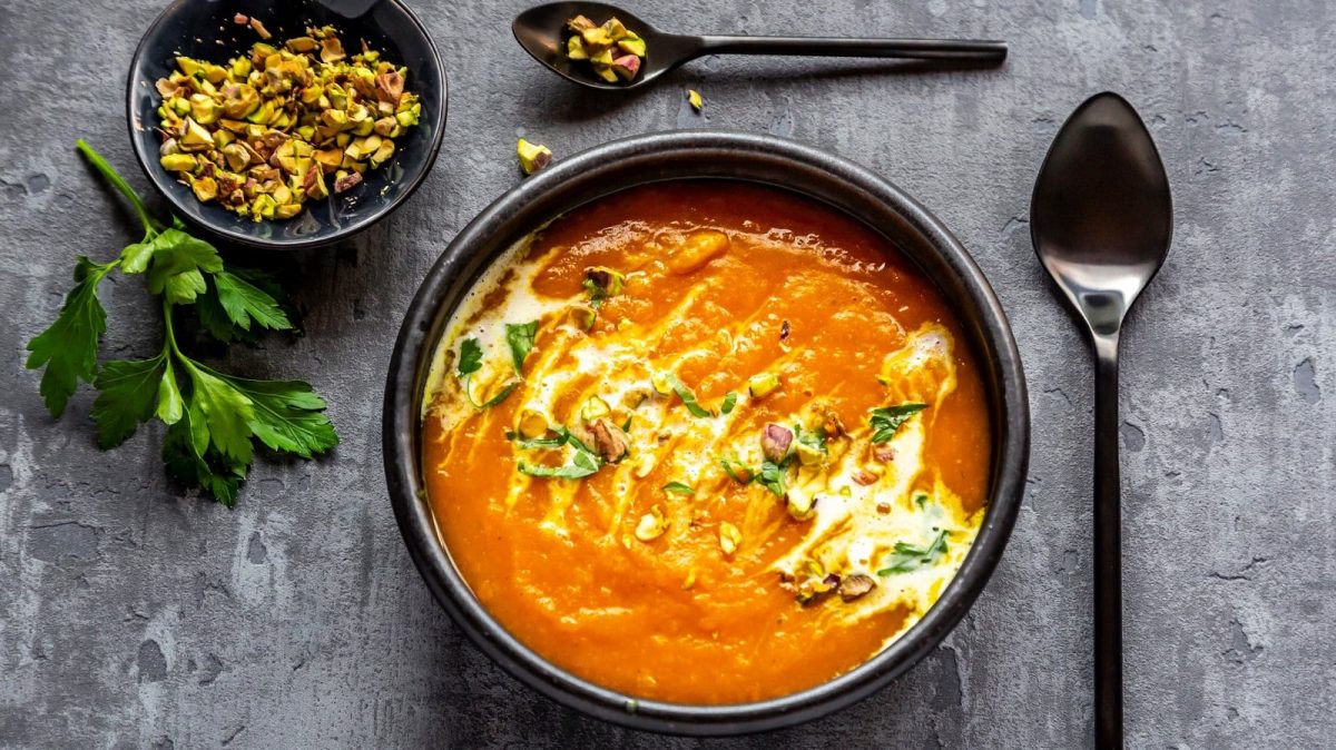 Eine Schüssel mit Süßkartoffelsuppe mit Chili und Pistazien gefüllt und Koriander garniert neben einem großen Lössel, einer kleinen Schale mit gehackten Pistazien und einem kleinen Löffel mit gehackten Pistazien vor einem grauen Hintergrund, von oben.