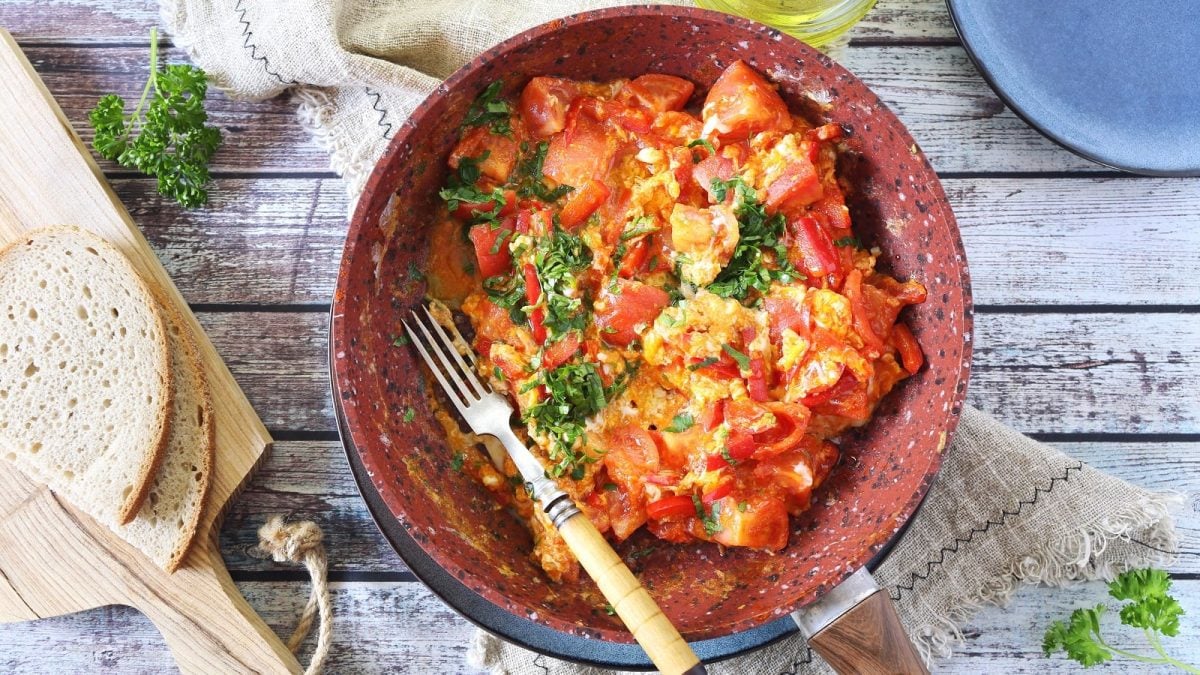 Eine Pfanne mit türkischen Menemen und einer Gabel neben einem Holzbrett mit geschnittenem Weißbrot, frischer Petersilie und einer Schüssel Olivenöl auf einem Holztisch, von oben.