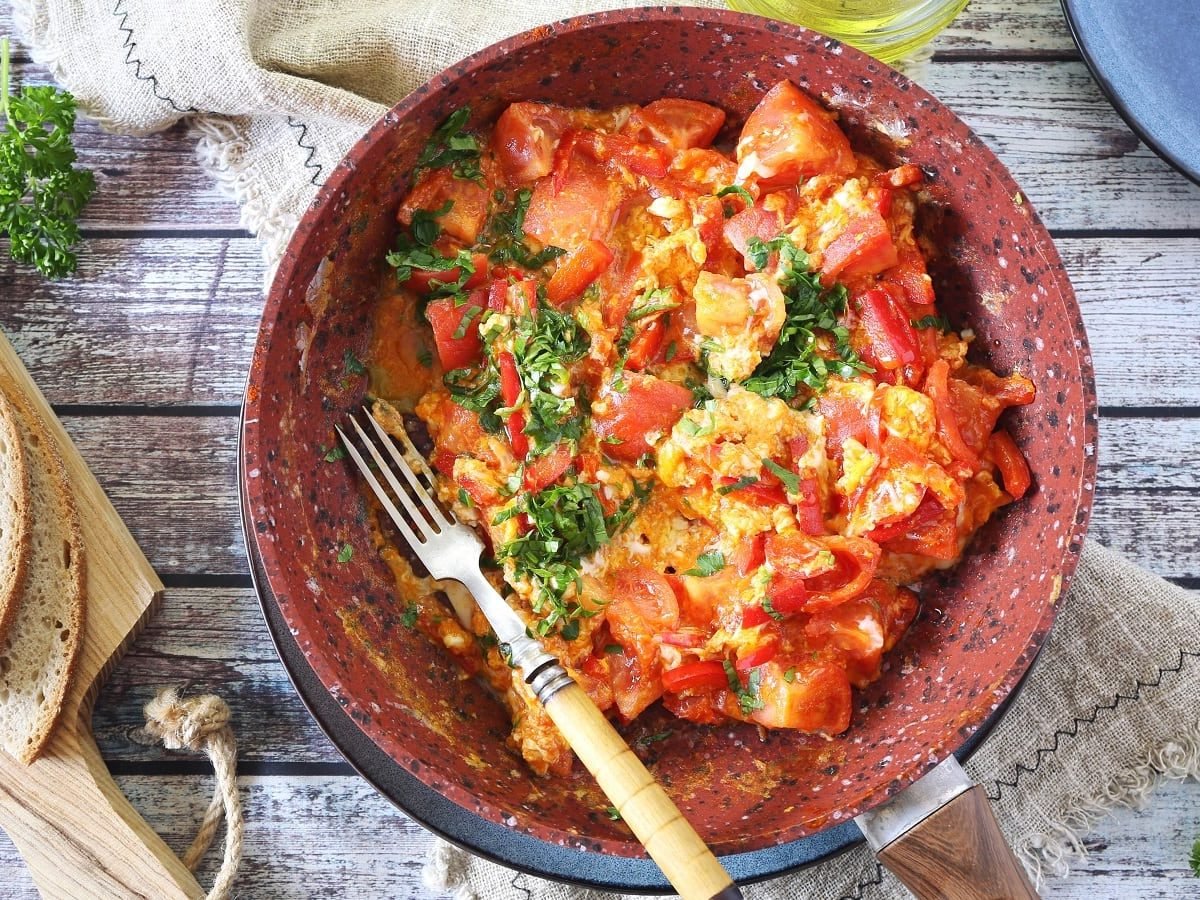 Eine Pfanne mit türkischen Menemen und einer Gabel neben einem Holzbrett mit geschnittenem Weißbrot, frischer Petersilie und einer Schüssel Olivenöl auf einem Holztisch, von oben.