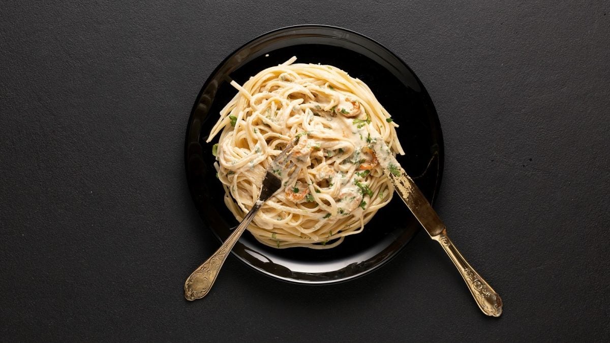 Ein schwarzer Teller mit einer Portion veganer Carbonara mit Pilzen und Messer und Gabel vor einem dunklen Hintergrund von oben.