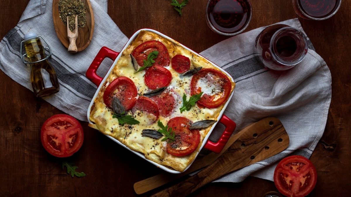 Eine Auflaufform mit veganer Lasagne, die mit Tomaten, veganem Käse, Basilikum und Rucola garniert ist neben einer Karaffe Wein, zwei gefüllten Gläsern, Gewürzen, frischen Tomaten und einem Holzheber, auf einem Küchentuch vor einem dunklen Holzhintergrund, von oben.