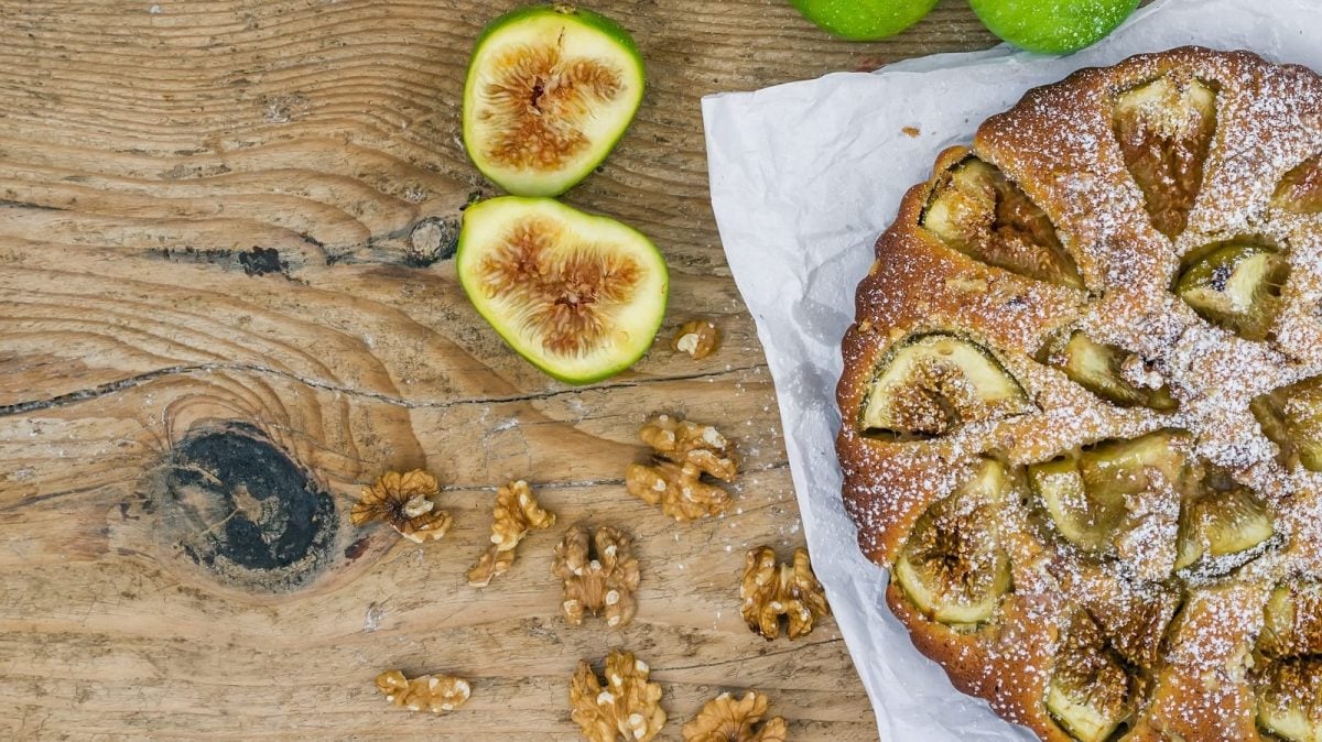Ein ganzer Walnuss-Feigenkuchen mit Puderzucker bestäubt auf einem weißen Backpapier neben aufgeschnittenen frischen Feigen und Walnusskernen vor einem Holzhintergrund, von oben.