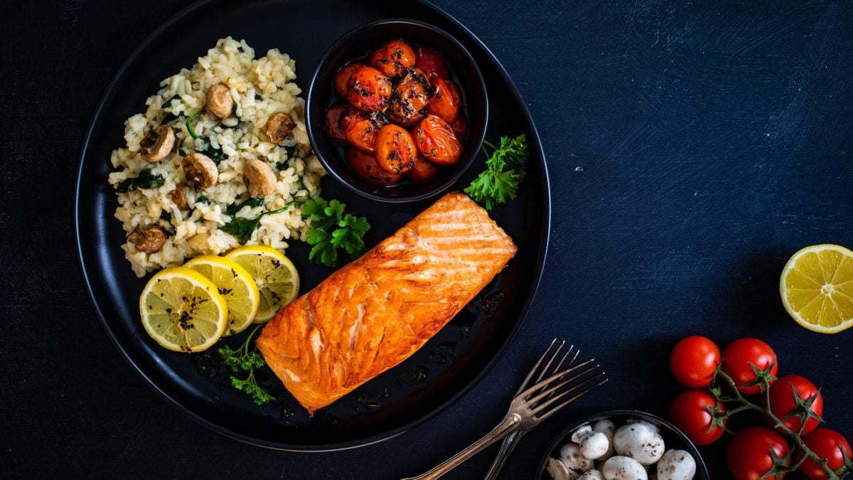 auf einem dunklen Untergrund steht ein dunkler Teller. Auf dem sieht man ein Zitronen-Risotto-mit-Lachs. Außerdem sind Tomaten und Pilze zu sehen.