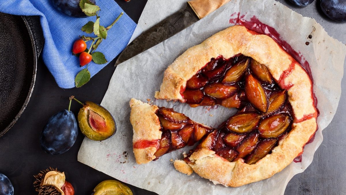 Eine angeschnittene Zwetschgen-Galette auf einem Backpapier neben einem Messer, aufgeschnittenen und ganzen Zwetschgen und einem blauen Küchentuch vor einem dunklen Hintergrund, von oben.