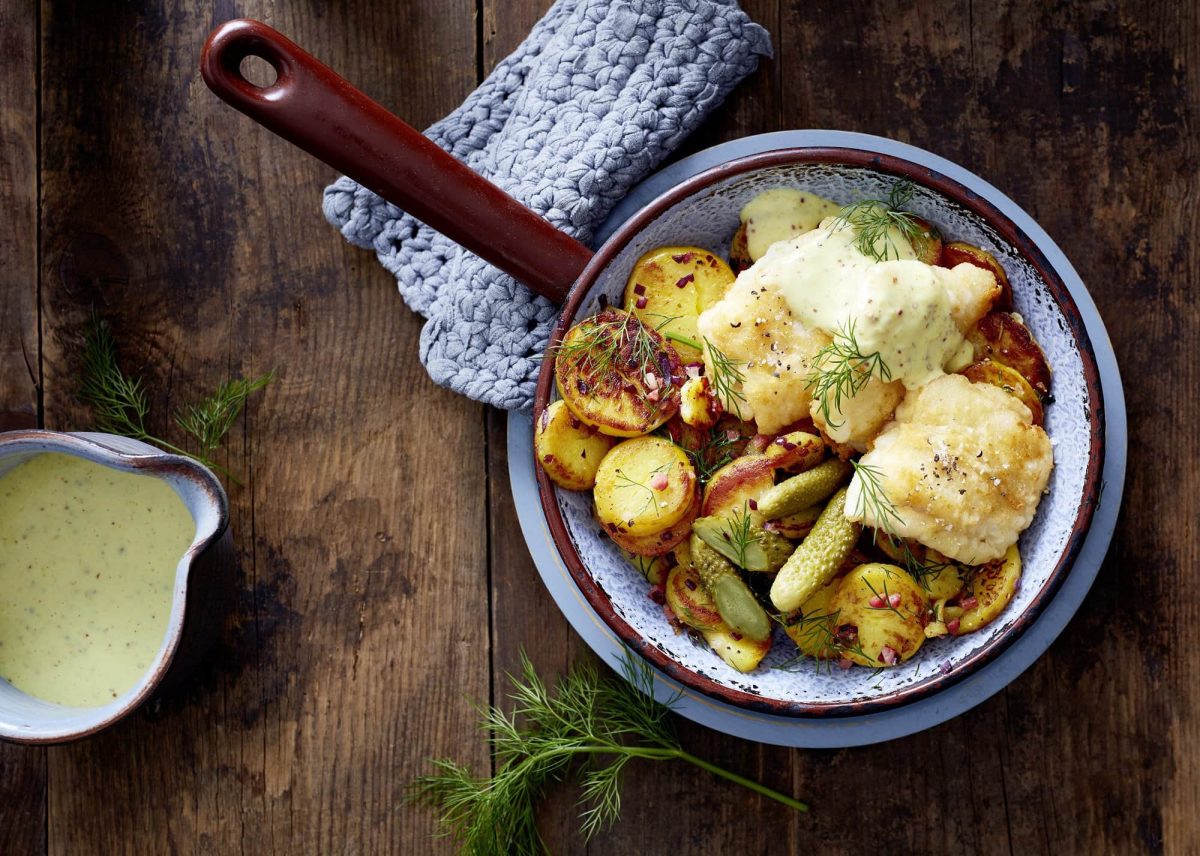 Pannfisch und Bratkartoffeln serviert in einer Pfanne. Die Senfsoße wird in einer Ausgusskanne dazu gereicht.