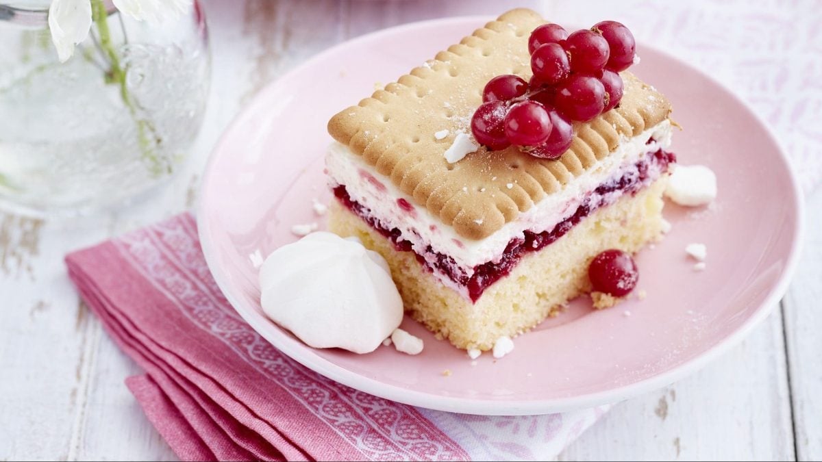 Ein Stück Butterkekskuchen mit Johannisbeeren angerichtet auf einem rosa Teller und mit rosa Serviette und Blumen als Deko im Hintergrund.
