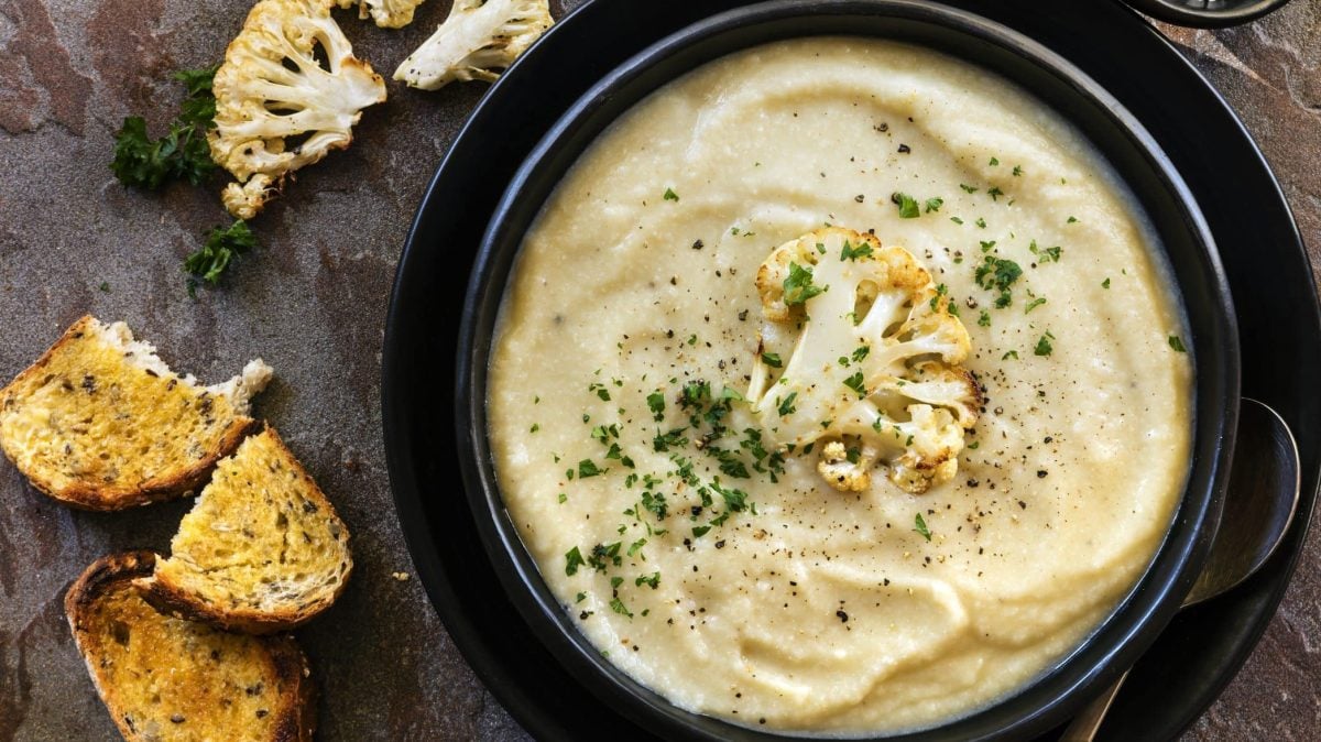 Die indische Blumenkohlsuppe füllt die schwarze Schüssel randvoll aus. Dazu gibt es etwas gerösteten Blumenkohlröschen als Garnitur und Röstbrot.