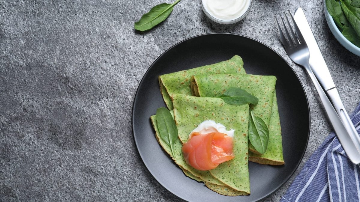 Drei Spinat-Pfannkuchen liegen zusammengeklappt auf einem schwarzen Teller. Dazu Crème fraîche und Lachs als Topping.