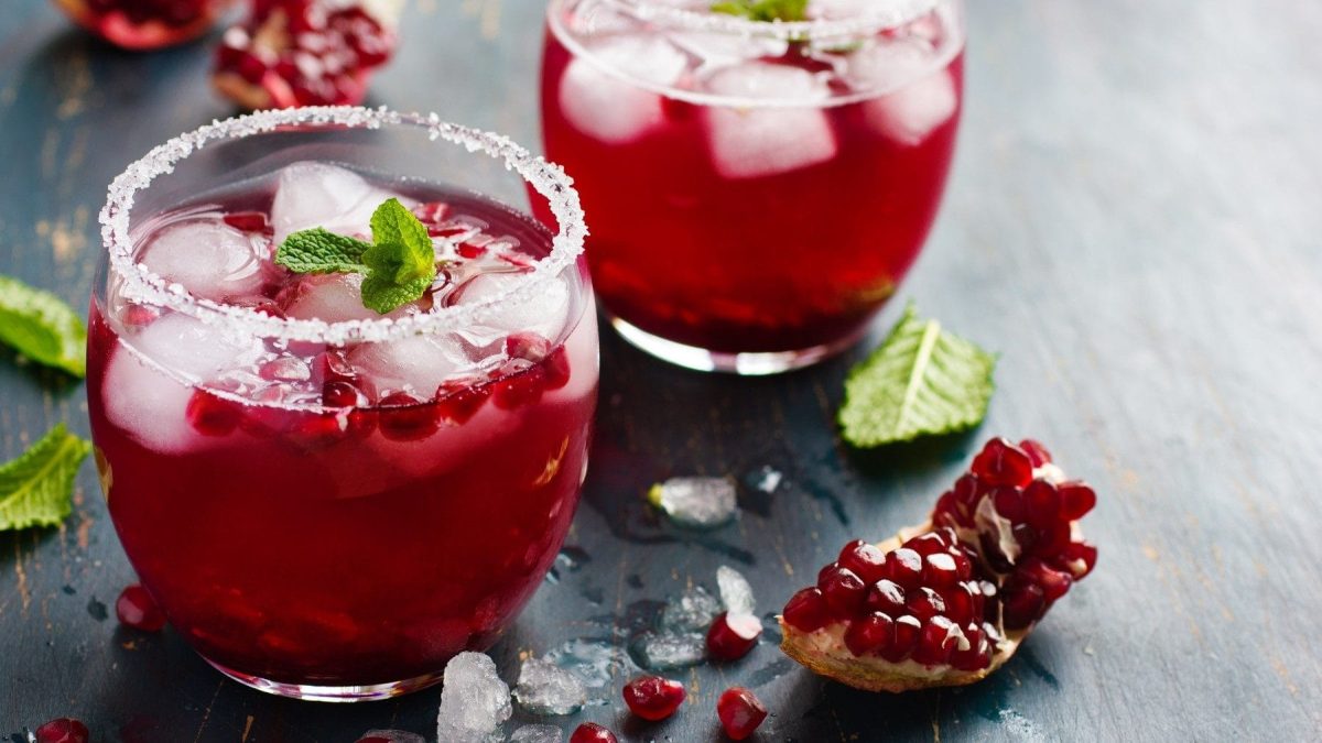 Zwei Gläser mit Zuckerrand und Granatapfel-Cocktail stehen auf einem dunklen Holztisch. Der Cocktail ist angerichtet mit Minze und Eis. Neben den Gläsern liegen Minzblätter und ein Stück Granatapfel.