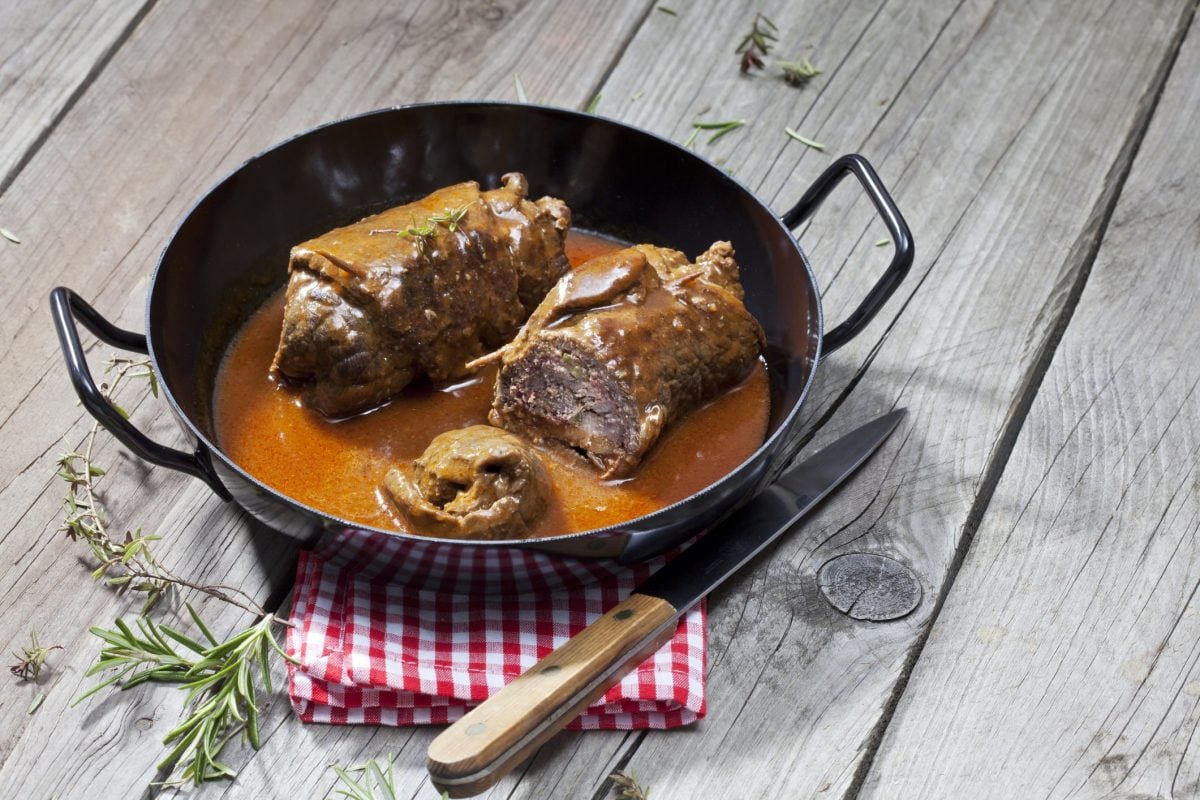 Rinderrouladen in gusseiserner Pfanne mit Soße auf grauem, hölzernem Untergrund.