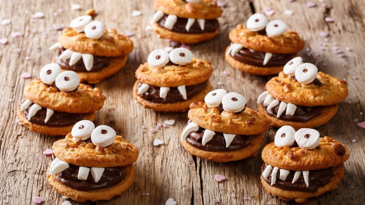 Mehrere Monster-kekse mit Marshmallow-Augen und Schokocreme liegen auf einem Holztisch. Um die Monster herum liegen Streusel in Herzform.