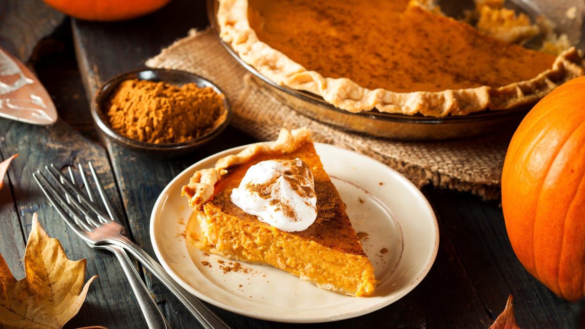 Angeschnittener Kürbiskuchen auf weißem Teller. Umgeben vom ganzen Kuchen und herbstlicher Dekoration.