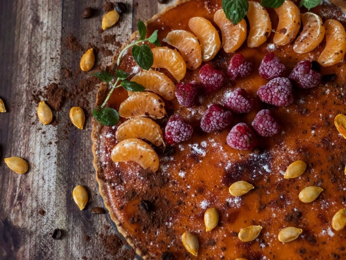 Ein Pumpkin Pie mit Obst ist angerichtet mit Mandarinen, Himbeeren, Kakaopulber und Puderzucker. Auf dem Kuchen und dem Untergrund liegen Kürbiskerne verteilt. Der Untergrund beseht aus braunem Holz.