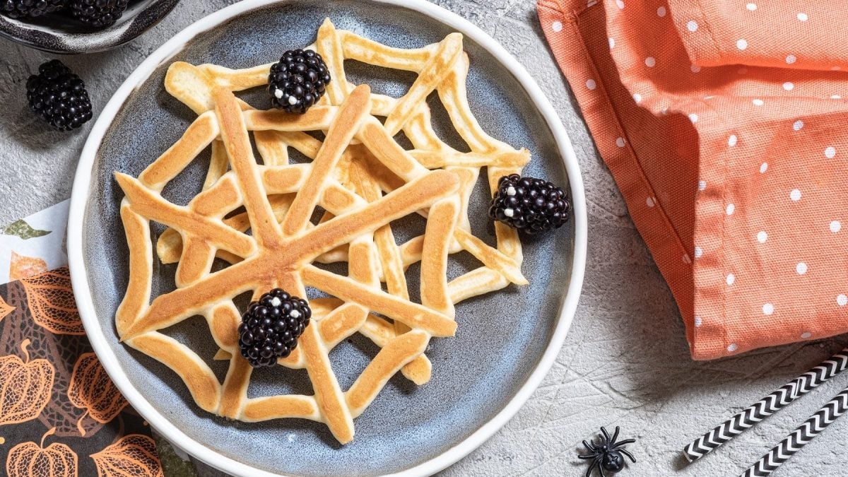 Zwei Spinnennetz-Pfannkuchen mit Brombeeren auf einem gemusterten Teller auf einem grauen Tisch. Um den Teller herum liegen Servietten, ein geschirrtuch, eine Schale mit Brombeeren und eine Plastik-Spinne. Außerdem liegen am rechten Bildrand zwei gemusterte Strohhalme.