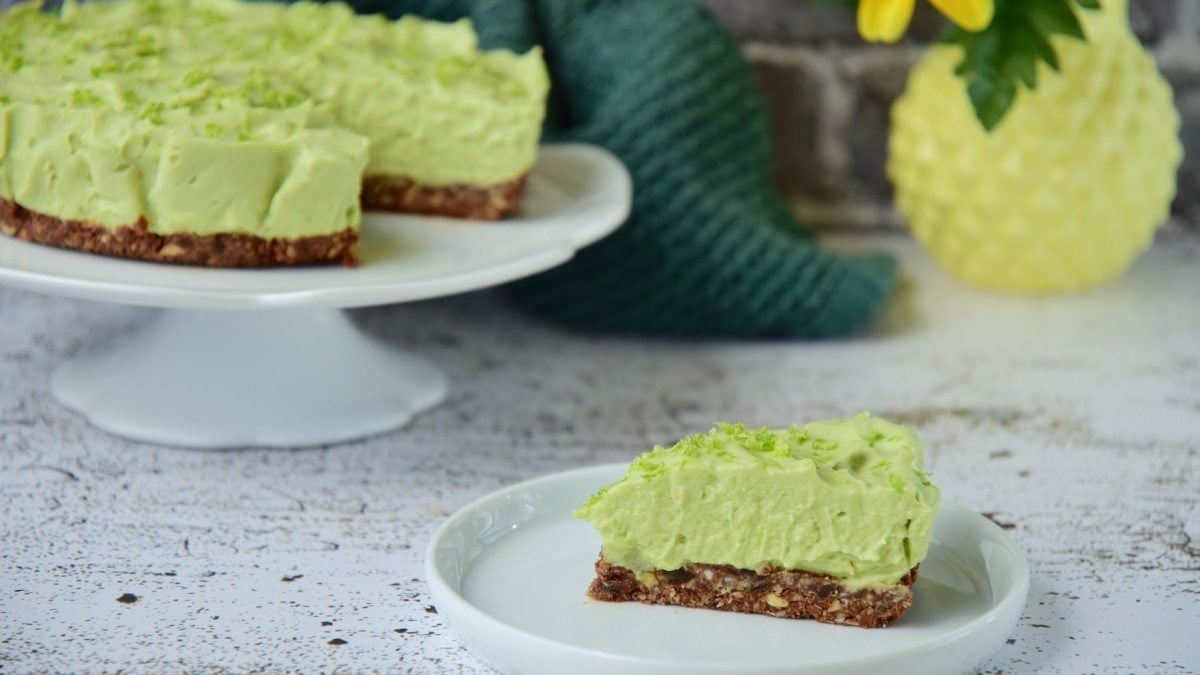 Ein Stück Avocado-Cheesecake mit Pistazien steht auf einem hellgrünen Teller auf einem gemusterten Untergrund. Im Hintergrund steht der restliche Kuchen auf einem Kuchentablett.