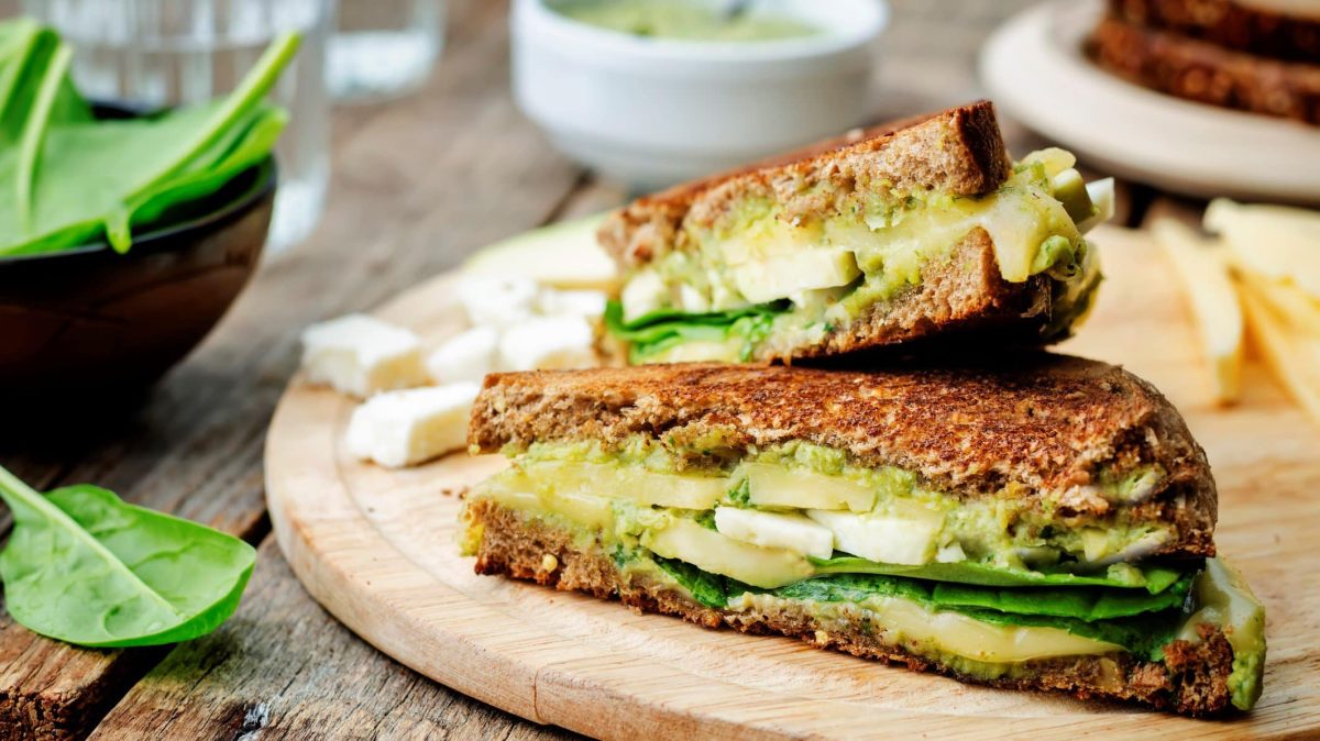 Auf einem Holzbrett liegen Avocado-Toasties, drumherum sieht man unscharf Zutaten wie Babyspinat, Käsewürfel oder Pesto.
