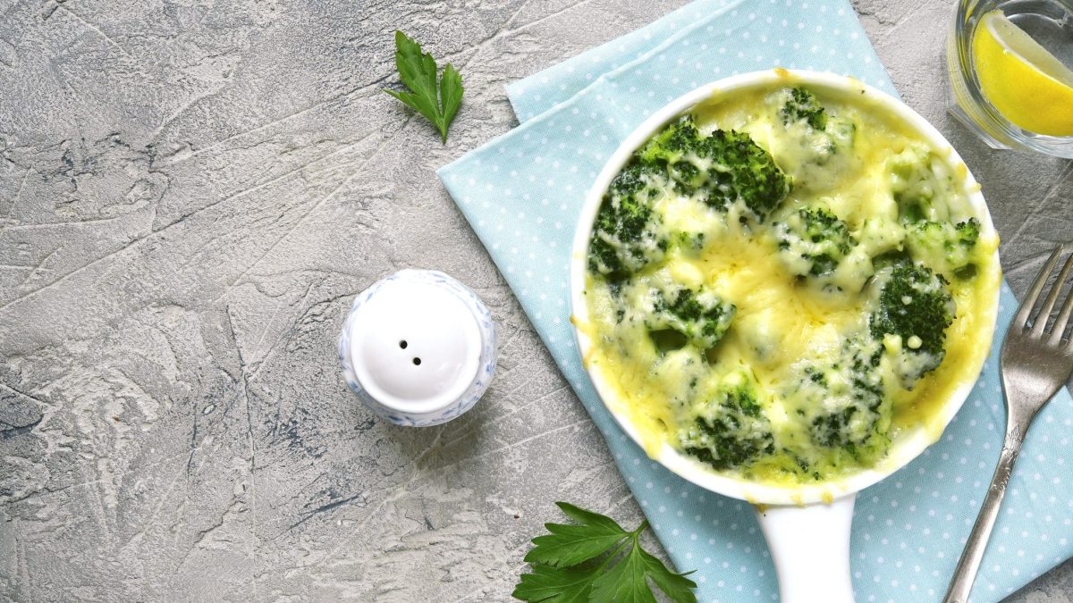 Das geschichtete Brokkoli-Blumenkohl-Gratin in einer Auflaufform. Dazu steht auf dem Tisch ein Salzstreuer, eine Gabel und ein Glas Wasser mit Zitrone.