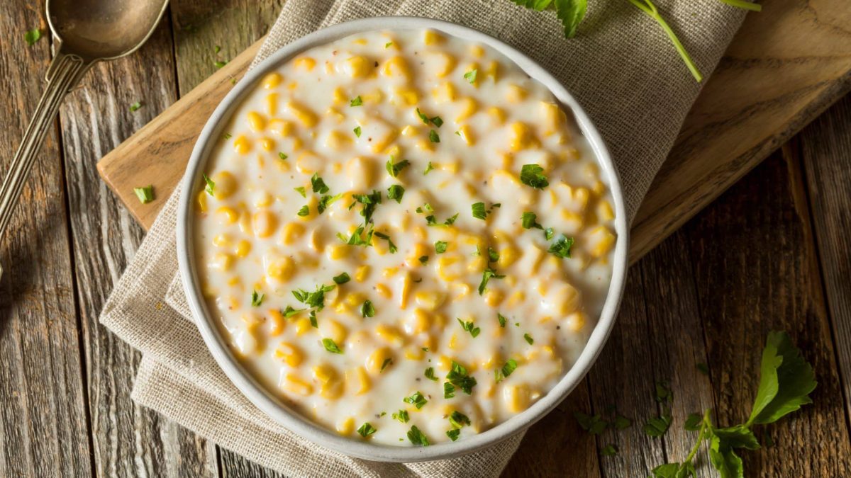 Eine Schale mit Creamed Corn, bestreut mit Petersilie, auf einem rustikalen Holztisch in der Draufsicht.