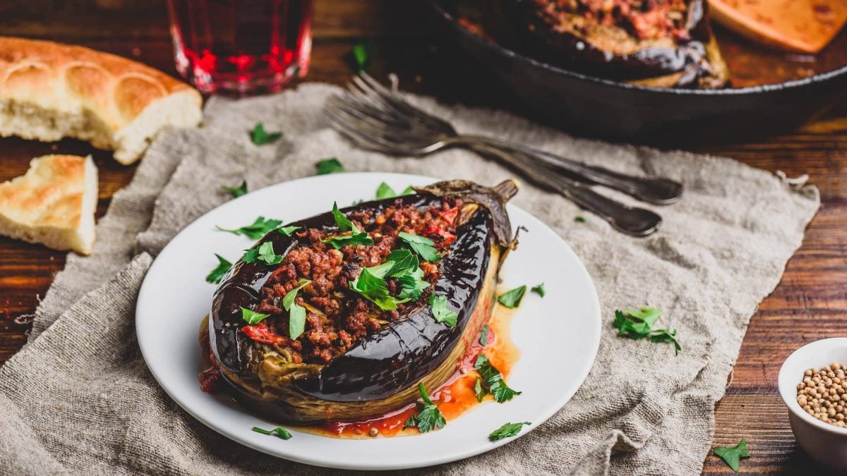 Auf einem weißen Teller liegt eine gefüllte Aubergine mit Nussmus, darüber gestreut ist Petersilie. Drumherum liegt etwas Brot, Besteck sowie eine Pfanne mit weiteren Auberginen.