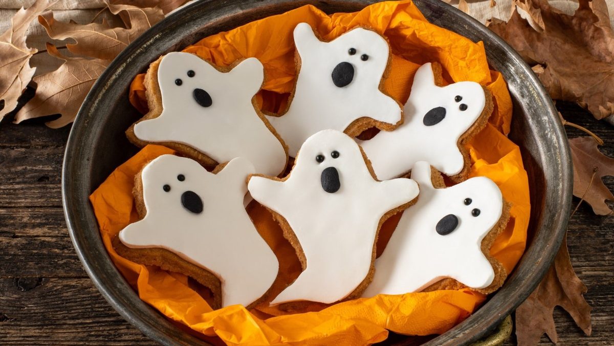 Sechs Geister-Kekse mit Fondant liegen in einer Schale mit orangener Serviette. Die Schüssel steht auf einem braunen Holztisch. Um die Schüssel herum liegen braune Laubblätter.