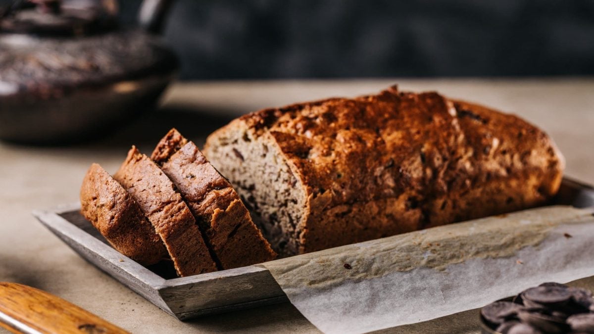 Ein angeschnittener Gewürzkuchen mit Datteln liegt auf einem Holztablett, im Hintergrund ist unscharf eine Teekanne zu erkennen.