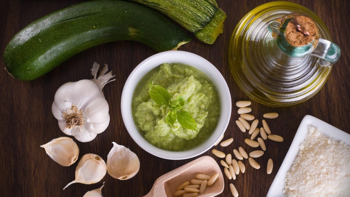 Auf einer Holzplatte sind zu sehen: eine Knoblauchknolle, eine Karaffe mit Olivenöl, zwei Zucchini, Pinienkerne, Salt, sowie eine runde Schale Zucchini-Pesto.