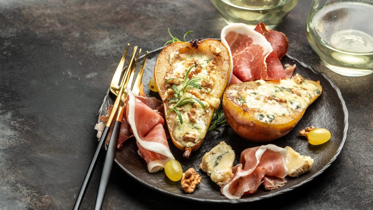 Auf einem rustikalen Tisch steht ein Teller, auf dem halbierte Birnen liegen. Diese sind mit Käse gefüllt. Neben den Birnenhälften liegen mehrere Scheiben Speck. Auf dem Teller liegen auch noch Walnüsse. Das Gericht heißt Birne mit Gorgonzola und Speck