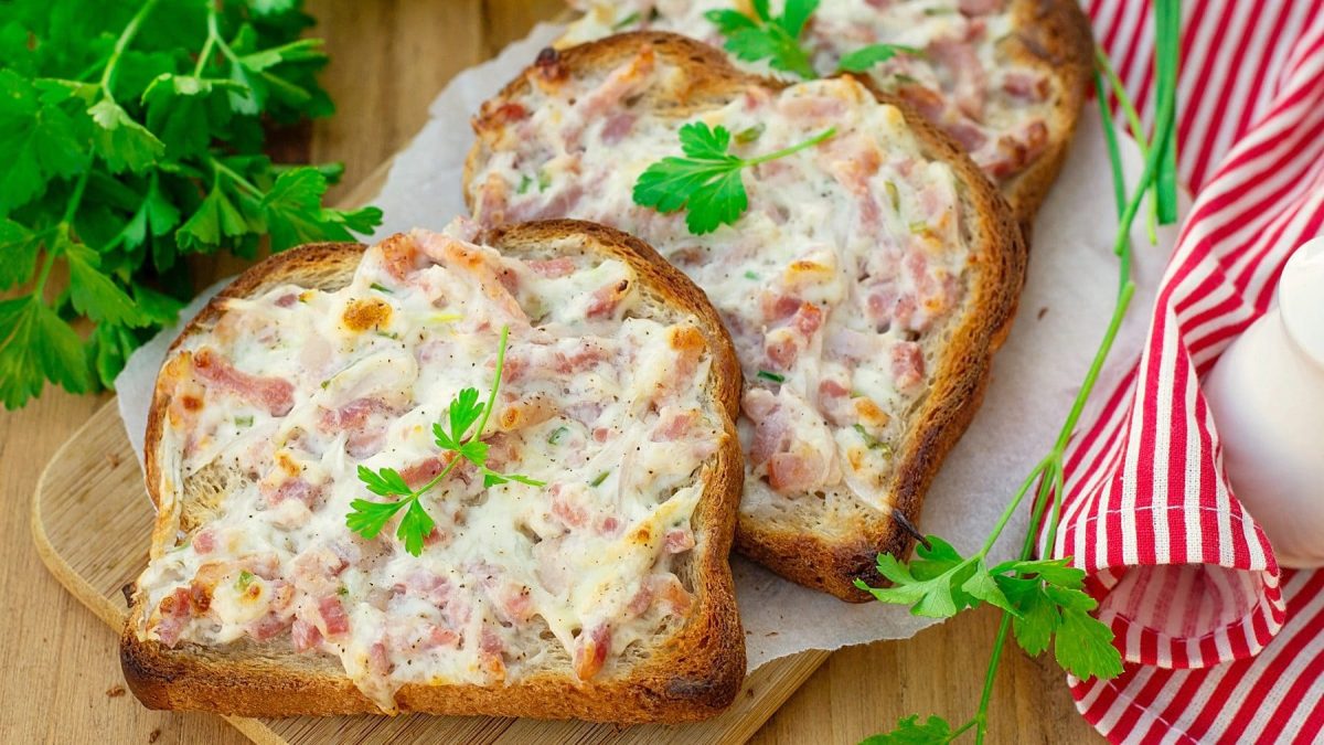 Drei Scheiben Toast sind mit einer Zwiebel-Speck-Mischung bestrichen. Sie liegen auf einem Papier und dann auf einem Holzbrettchen. Links ist Petersilie und rechts ein rot-weiß-gestreiftes Handtuch.