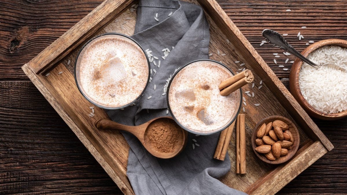 In der Draufsicht: ein Holztablett, auf dem zwei Gläser Horchata mit Eis und Zimtstangen stehen. Drumherum sind Zutaten wir Zimt, Mandeln und Reis verteilt.