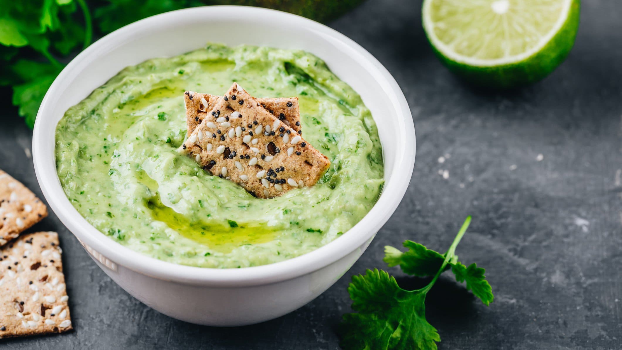 Eine weiße Schale mit Hummus mit Koriander, darin stecken ein paar Cracker. Daneben sieht man frischen Koriander sowie eine aufgeschnittene Limette.