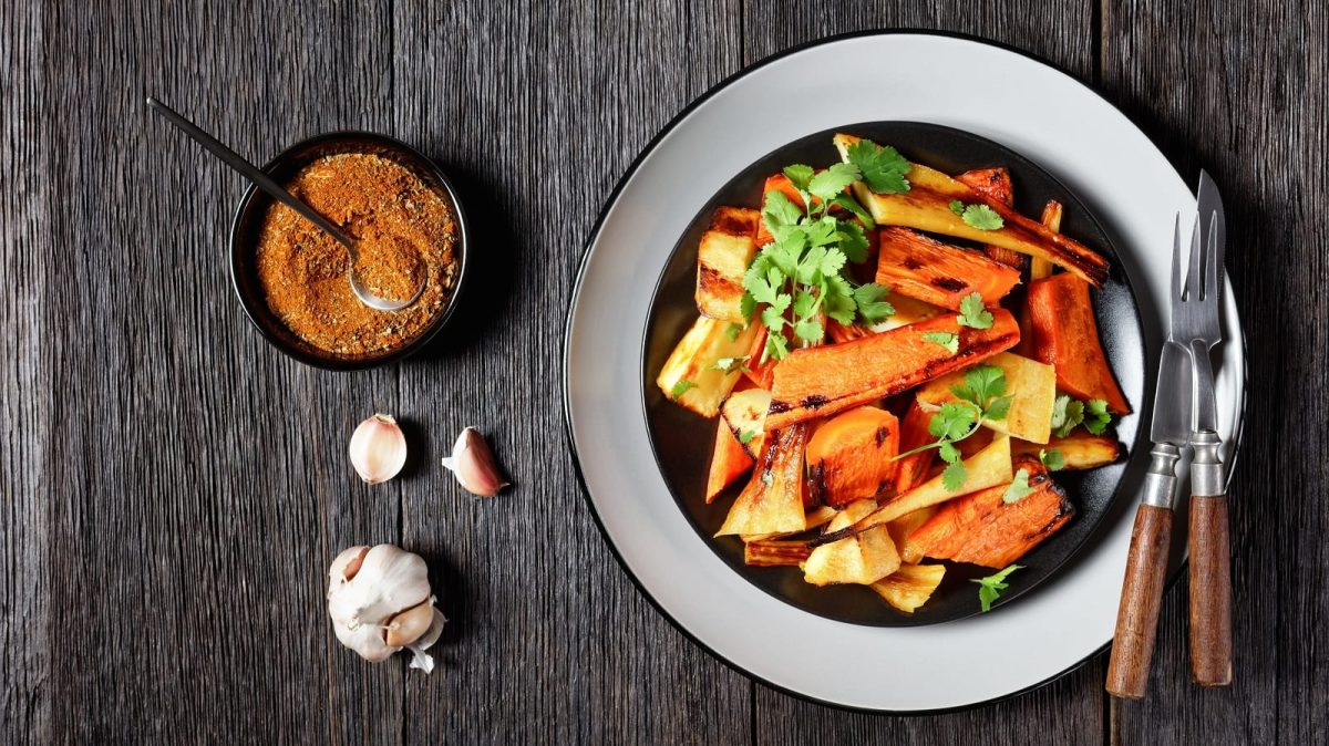 Ein Teller gefüllt mit gebackenen Karotten und Pastinaken, garniert mit frischem Koriander, neben einer Knoblauch-Knolle, einer kleinen Schüssel mit marokkanischen Gewürzen und Messer und Gabel, vor einem Holzhintergrund, von oben.