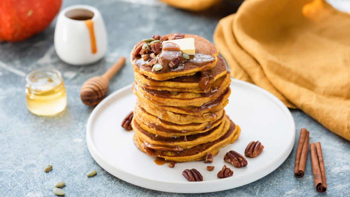 Ein weißer Teller mit gestapelten Kürbis-Pancakes. Darauf Pecannüsse, ein Stück Butter und etwas Sirup. Drumherum sieht man ein Honigglas, Zimtstangen und ein Stück von einem Kürbis.