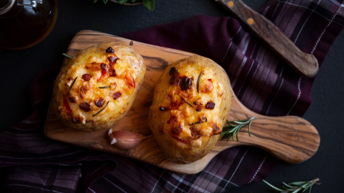 In der Draufsicht sieht man zwei gefüllte Kumpir mit Apfel, Paprika und viel Käse, die auf einem Küchenbrett liegen. Dunkler Untergrund mit einem Geschirrtuch.