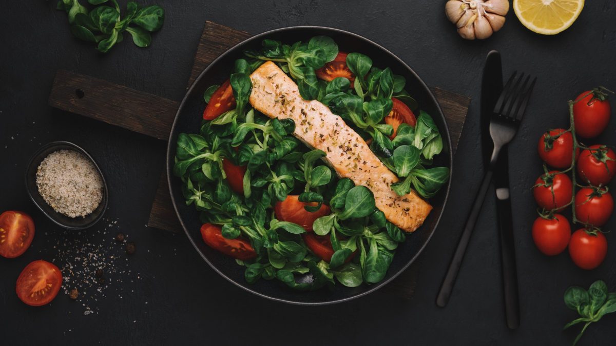 Ein Teller mit Lachs mit Feldsalat und Tomaten auf dunklem Untergrund in der Draufsicht. Drumherum sieht man einzelne Zutaten wie Tomaten, Salz oder eine halbe Zitrone, sowie Besteck.
