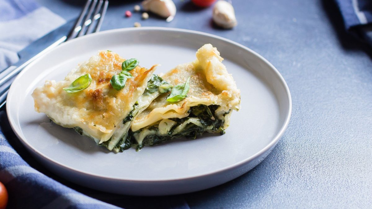 Ein Teller mit zwei Stück Lachs-Spinat-Lasagne bestreut mit frischem Basilikum. Im Hintergrund sieht man unscharf ein paar Knoblauchzehen, Pfefferkörner, Besteck und Geschirrtücher.