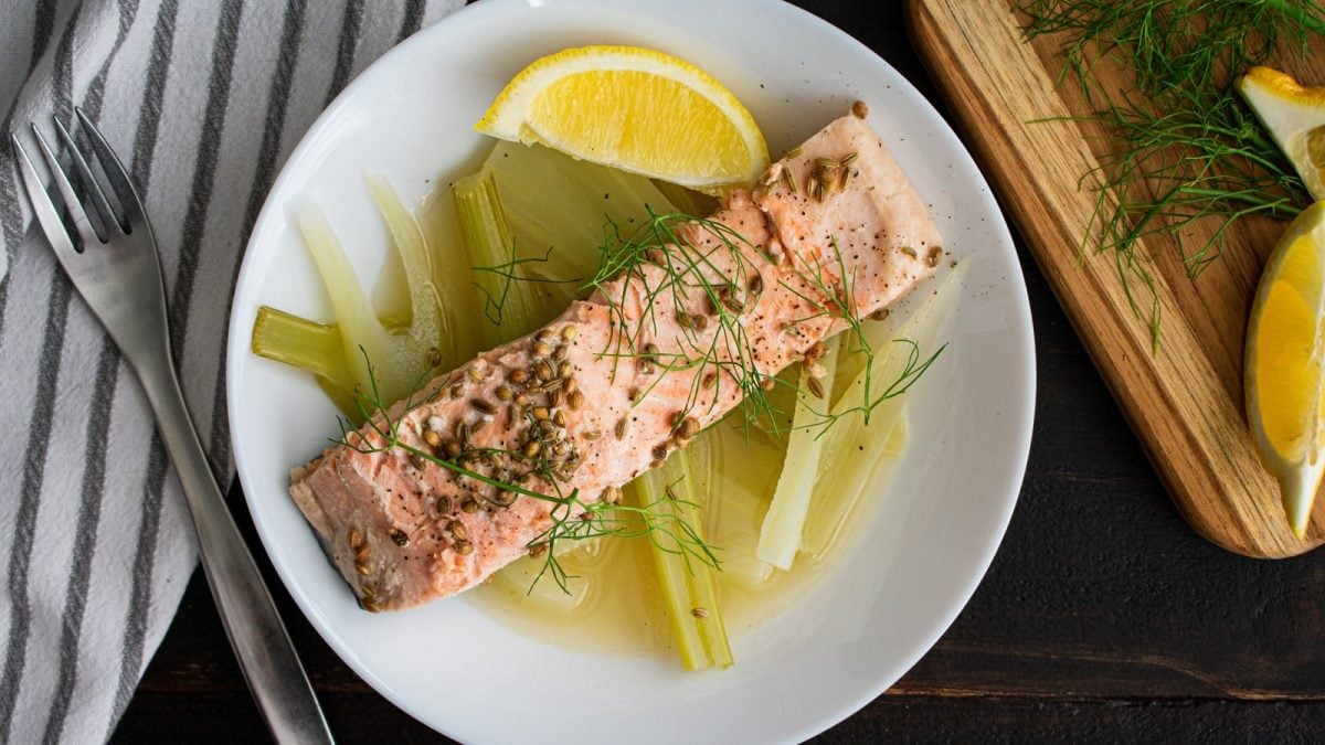 Ein Stück gebackenes Lachsfilet auf Fenchel-Zitronenbett mit Fenchelsamen, Fenchelgrün und frischer Zitrone serviert neben einer Gabel, einem Holzbrett mit Zitronen und Fenchel und einem gestreiften Küchentuch, vor dunklem Hintergrund, von oben.