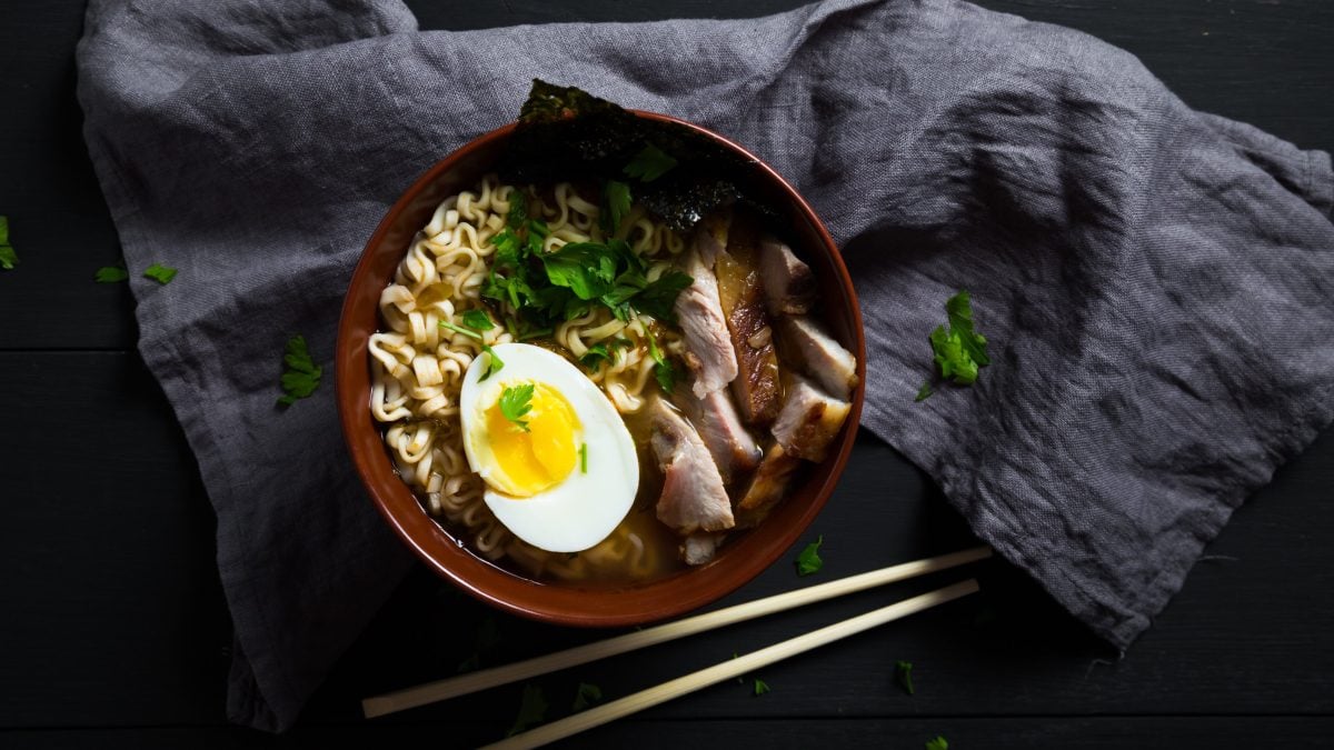 Eine Schüssel mit Miso Ramen mit eingelegtem Schweinebauch, einem halbem Ei, frischem Koriander und einem Noriblatt als Topping, neben einem Paar Essstäbchen, auf einem grauen Küchentuch, vor dunklem Hintergrund, von oben.