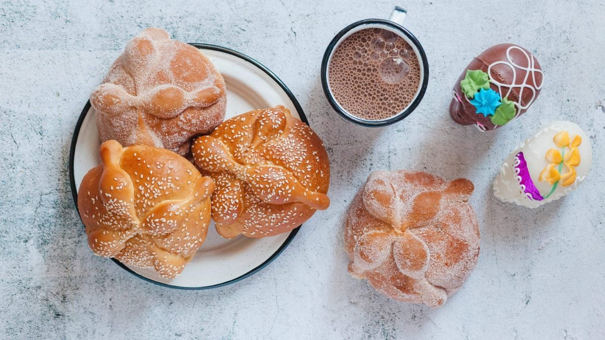 Ein Teller mit drei Pan de Muerto bestreut mit Zucker und Sesam neben einer Tasse heißer Schokolade, einem weiteren Brötchen und zwei Schokoladen Totenköpfen.