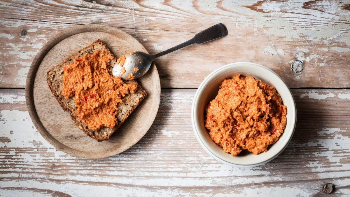 In der Draufsicht sieht man eine Schale mit Paprikafrischkäse, daneben steht ein Teller mit einer Scheibe Brot, die mit dem Frischkäse bestrichen ist. Alles auf Holz.