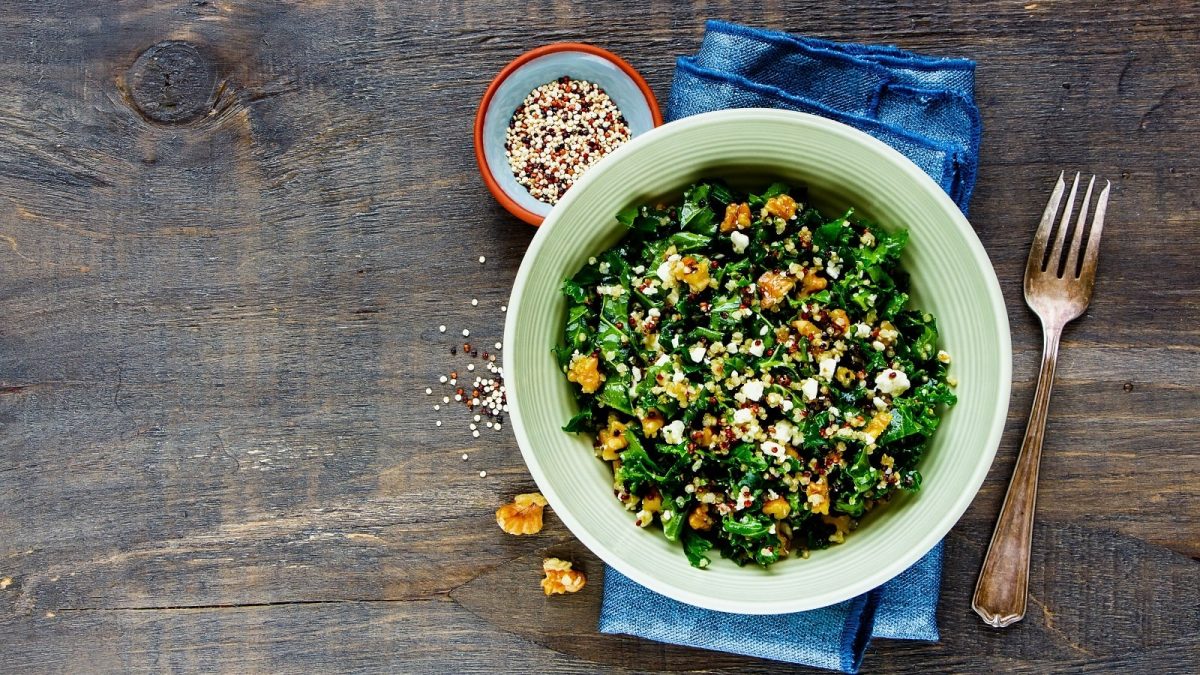 Eine hellgrüne Schüssel gefüllt mit dem Quinoa-Grünkohl-Salat garniert mit gerösteten Walnüssen und zerbröseltem Feta-Käse auf einem blauen Küchentuch neben einer kleinen Schüssel rohen Quinos-Körnern, zwei Walnüssstücken und einer Gabel vor einem Holzhintergrund, von oben.