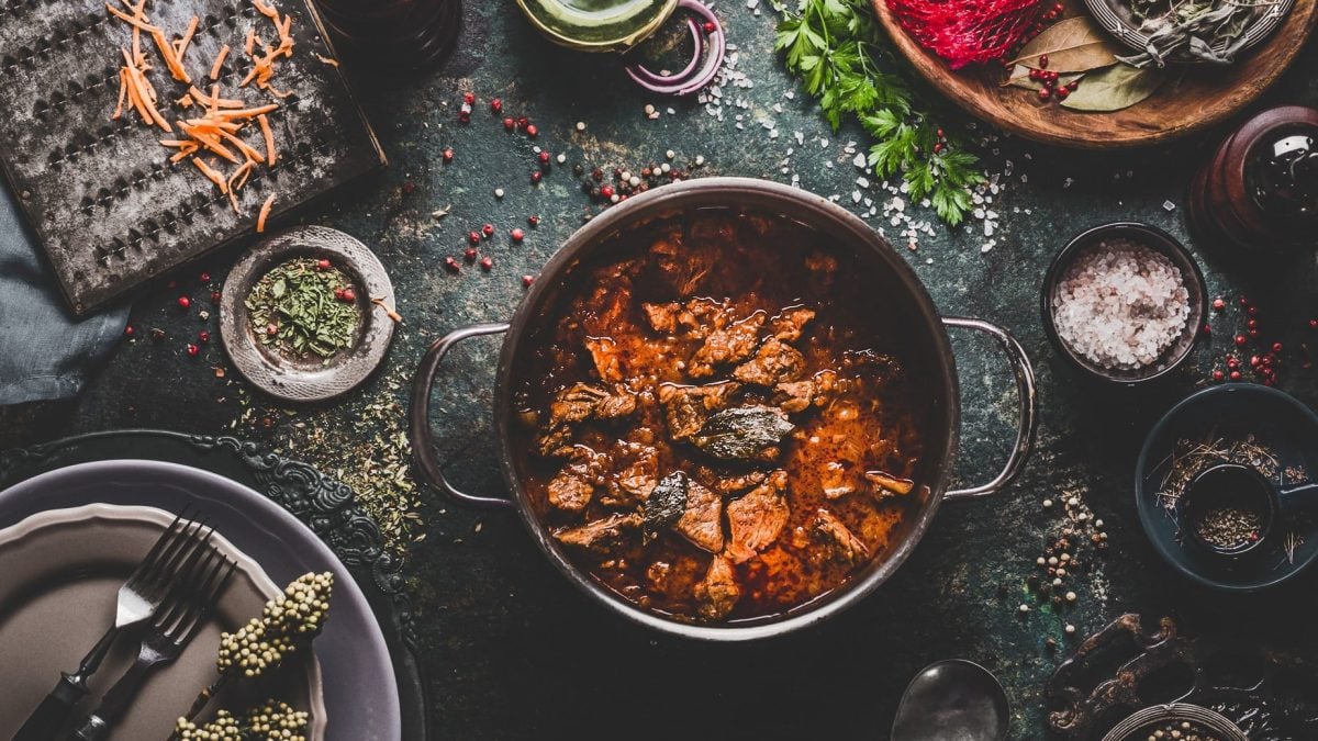 Ein Topf mit Rindergulasch mit Coa in der Draufsicht auf dunklem Untergrund. Drumherum sind eine Vielzahl von Schälchen mit Zutaten zu sehen.