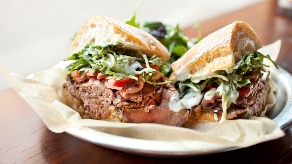 Ein halbiertes Roastbeef-Sandwich mit Rucola auf einem Teller, der auf einem Holztisch steht.