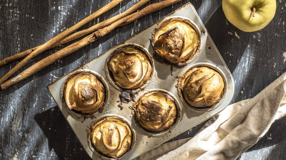 Auf einem dunklen Hintergrund ist eine Muffinform mit sechs saftigen Apfel-Muffins. Dabene liegen Zimtstangen und ein Apfel.