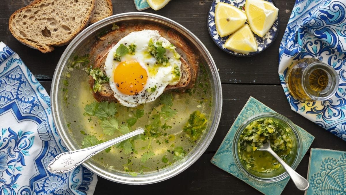 Eine Schüssel Sopa Alentejana neben einer kleinen Glasschüssel mit einer Knoblauch-Koriander-Paste, einer Karaffe mit Olivenöl, einem Teller mit Zitronenstücken und zwei Scheiben Brot, vor einem Holz-Hintergrund, von oben.