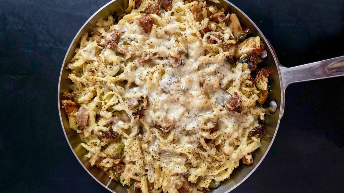Eine große Portion Spätzle-Rosenkohl-Pfanne mit Bacon und Käse in einer Pfanne präsentiert vor dunklem Hintergrund, von oben.