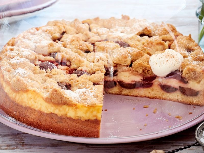 Ein angeschnittener Streuselkuchen mit Pflaumen und Zimt auf einem lila Teller bedeckt mit einer Glas-Glocke neben einer kleinen Schüssel Puderzucker vor einem hellen Hintergrund.