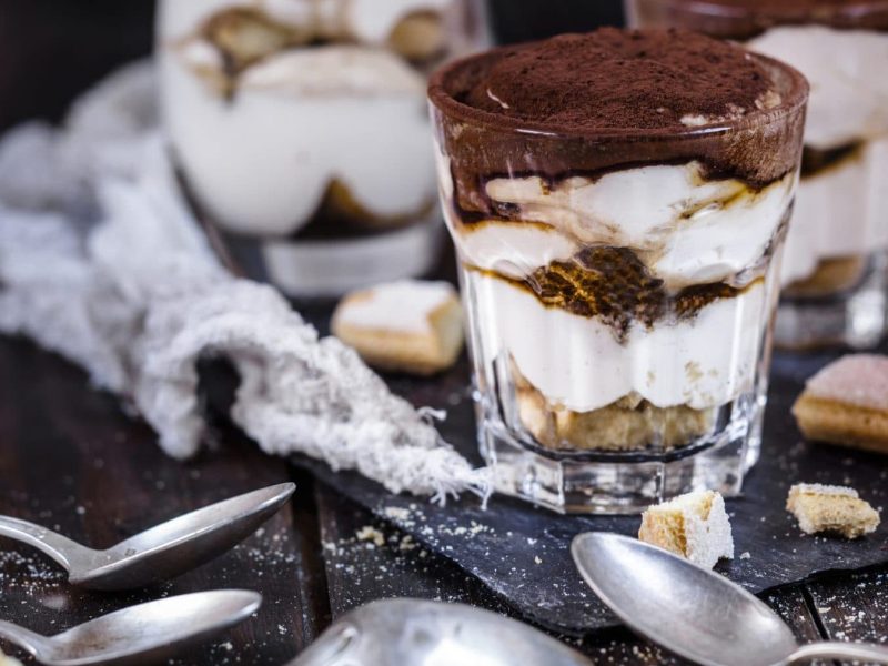 In kleinen Glaesern ist das italienische Dessert Tiramisu angerichtet. Daneben lieben vier Loeffel sowie einige Biskuits-